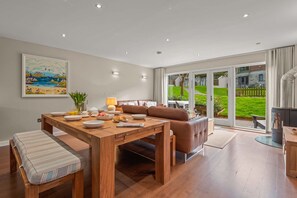 Ein modernes Wohnzimmer mit einem Holzesstisch, einer Couch und einem großen Fenster mit Blick auf einen grünen Garten und Nachbarhäuser.