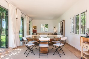 A spacious dining area with a round table and chairs, a sofa, and a view of greenery through large windows. - Es Sestador (Porto Cristo) - 784526 Es Sestador (Manacor)