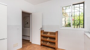 Een kamer met betegelde vloer, een raam met uitzicht op groen, een houten plankunit en een deur naar een andere kamer.