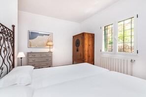 A bedroom with a bed, a wooden cabinet, a nightstand with a lamp, a framed picture on the wall, and a window with a view of trees.
