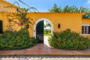A yellow building with a black door, surrounded by greenery and a tiled pathway. - Es Sestador (Porto Cristo) - 784526 Es Sestador (Manacor)