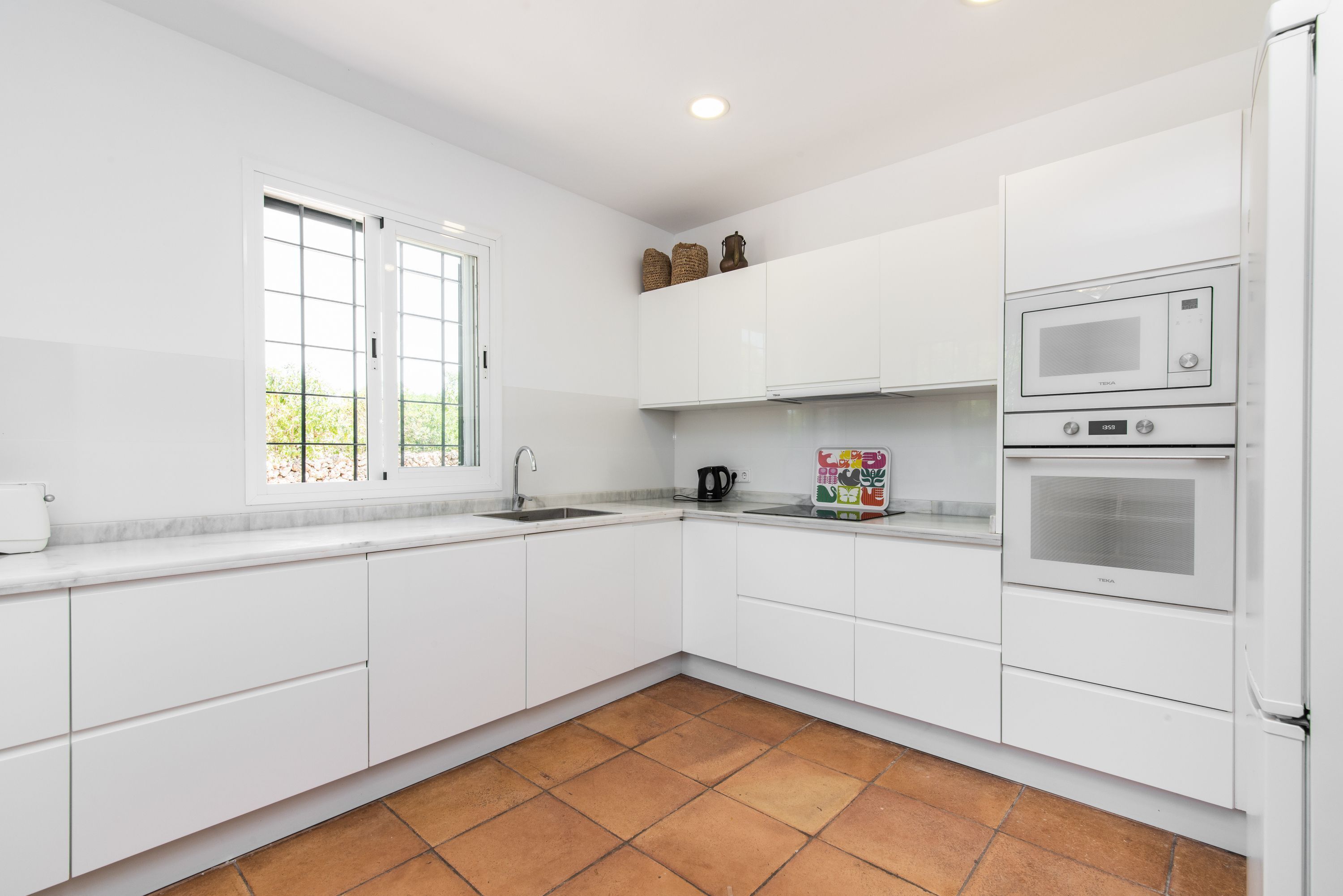 A modern kitchen with white cabinets, a double oven, a microwave, and a window with a view of greenery.