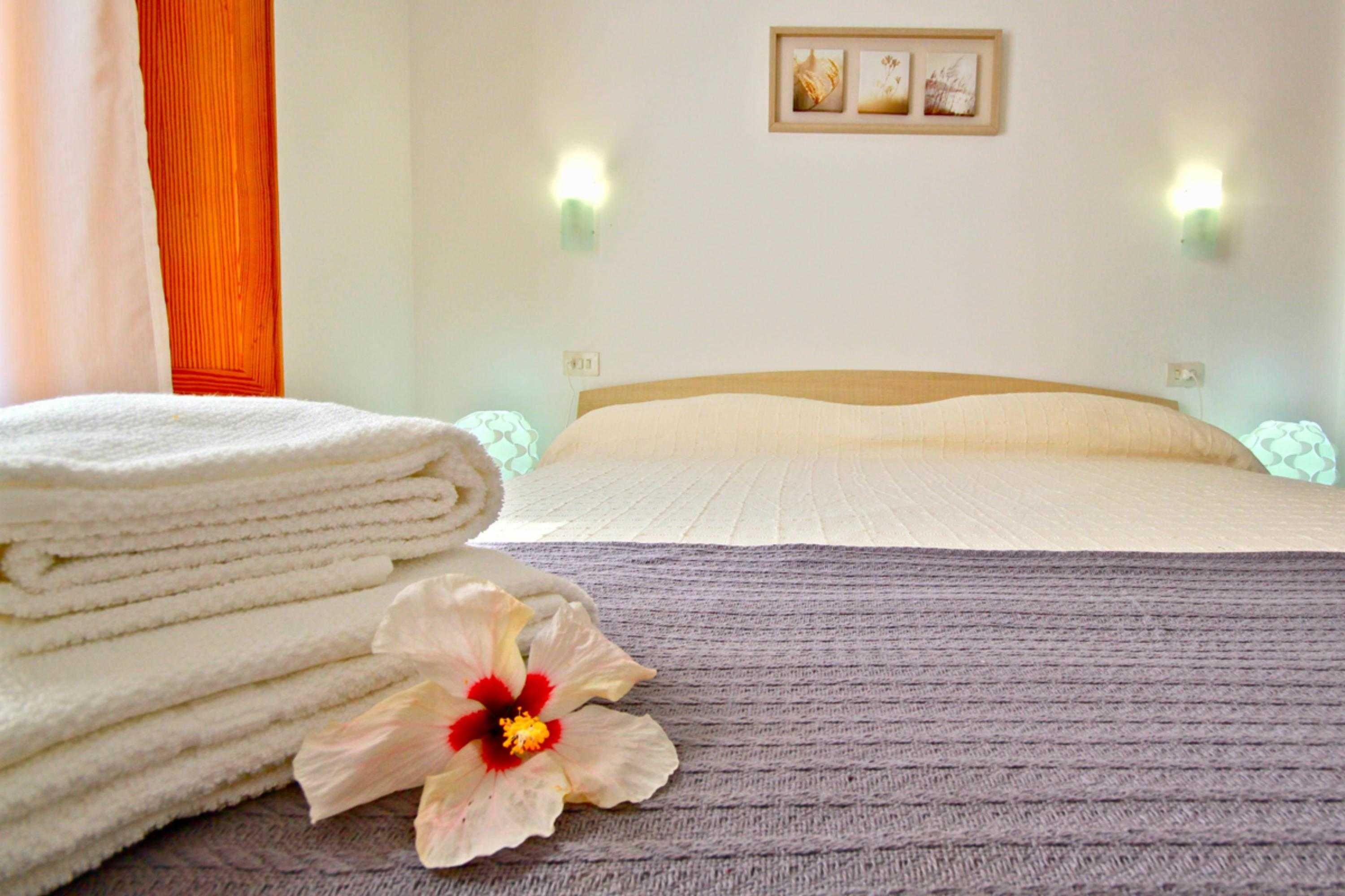 A neatly made bed with a folded white towel and a single hibiscus flower on a textured gray bedspread.