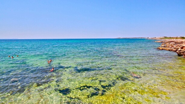 Una playa con agua cristalina, costa rocosa y gente nadando.