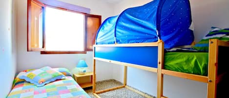 Una cama litera con dosel azul, una mesita de noche pequeña con una lámpara, una ventana con vistas y una colcha colorida en la cama inferior.