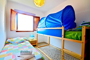 Una cama litera con dosel azul, una mesita de noche pequeña con una lámpara, una ventana con vistas y una colcha colorida en la cama inferior.