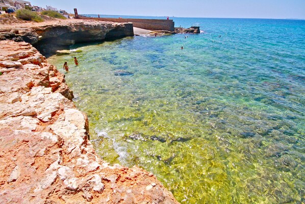 A coastal rocky area with clear water, a pier, and a distant view of the sea.