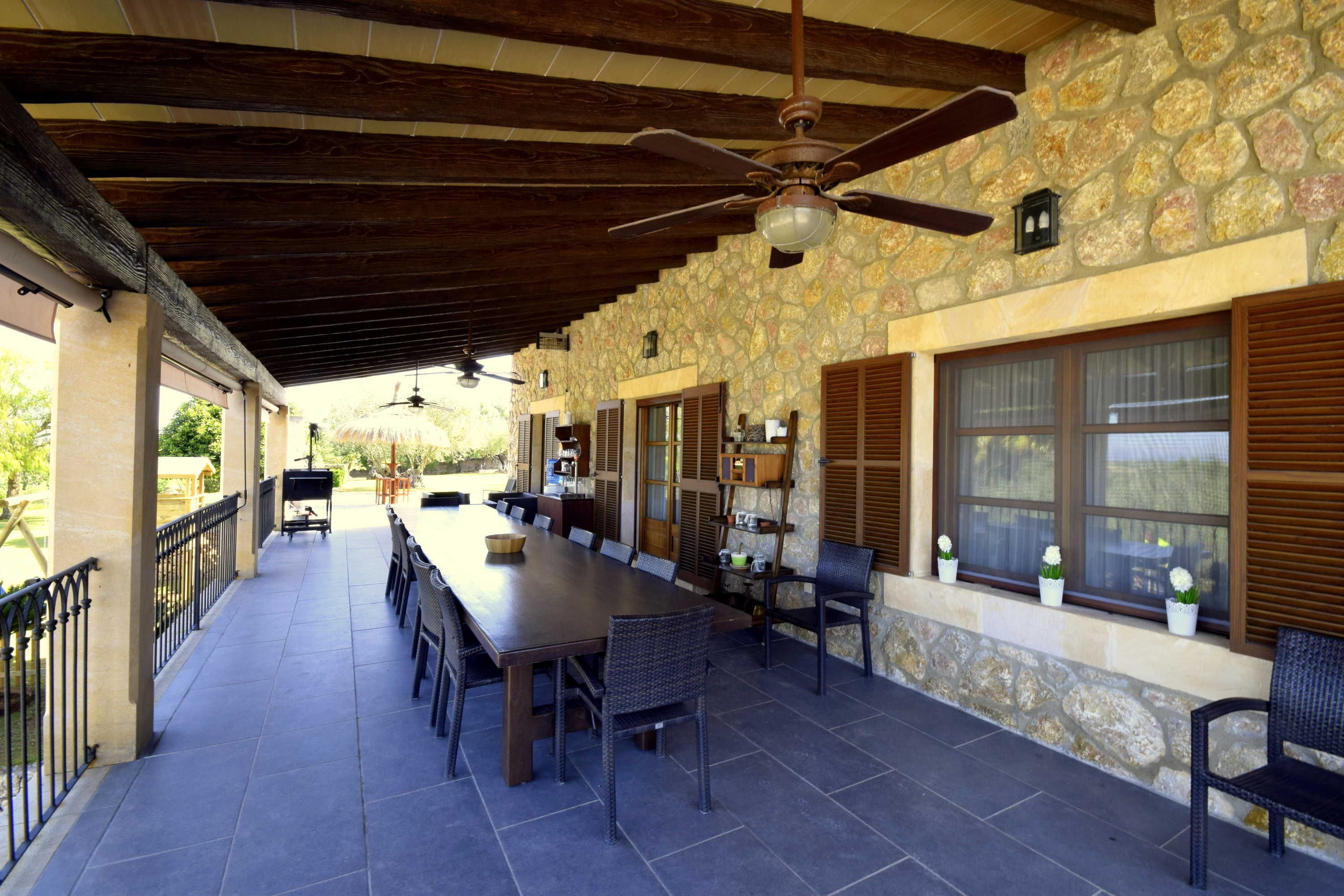 Un patio exterior con una mesa larga y sillas, paredes de piedra, vigas de madera y un ventilador de techo.