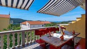 Un balcone con un tavolo apparecchiato per mangiare, affaccia su una cittadina costiera con edifici dai tetti rossi e un cielo azzurro limpido.