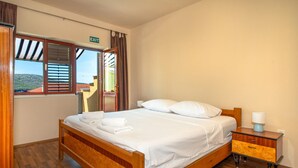 Una camera d'albergo con un letto in legno, lenzuola bianche, un comodino con una lampada e un segnale di uscita sopra una porta.