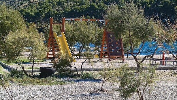 Un'area giochi con uno scivolo giallo e strutture per arrampicarsi, circondata da alberi e un corpo d'acqua sullo sfondo.