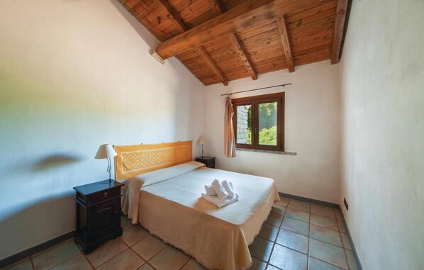 Un dormitorio con techo de madera, una cama con cabecera, una mesita de noche, un ventanal con vista a follaje y suelo de baldosas.