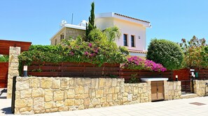 A two-story house with a stone wall in the foreground, a wooden fence, and a variety of flowering plants. - Private Detached Villa With Pool Ideal for Families (Coral Bay)
