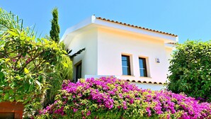A white building with two windows and a small balcony, surrounded by lush greenery and vibrant pink flowers. - Villa Olivia (Coral Bay)