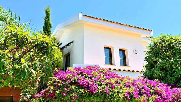 A white building with two windows and a small balcony, surrounded by lush greenery and vibrant pink flowers. - Private Detached Villa With Pool Ideal for Families (Coral Bay)