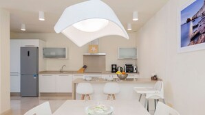 Eine moderne Küche mit weißen Schränken, einem Kühlschrank, einer Mikrowelle und einer Kaffeemaschine. Ein Esstisch mit Stühlen und eine Schüssel Obst sind ebenfalls vorhanden.