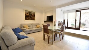Una sala de estar con un sofá, una mesa de comedor con sillas, un televisor montado en la pared y un ventanal con vistas.