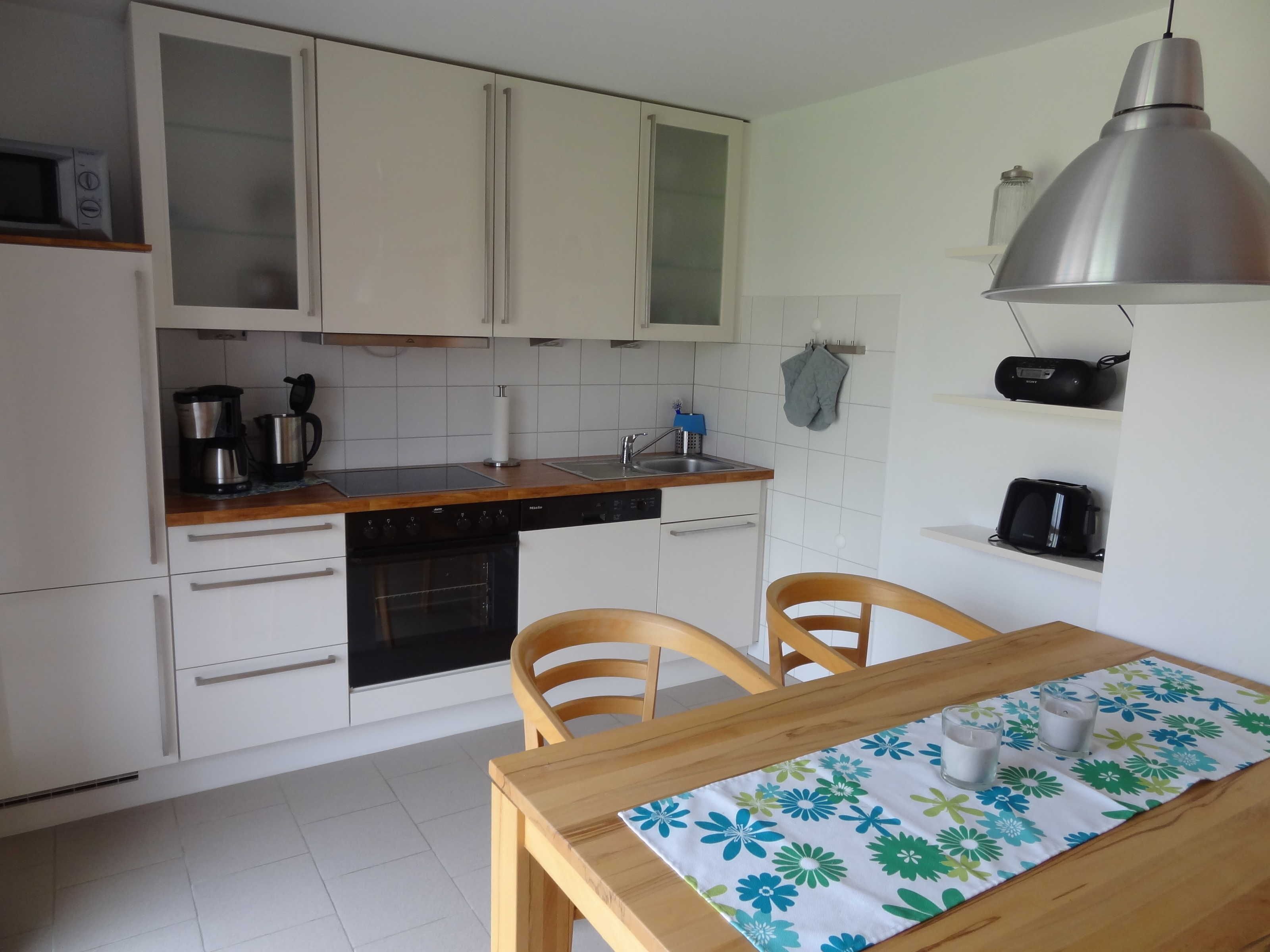 Una cocina compacta con armarios blancos, horno empotrado, microondas y lavamanos. Hay una mesa de comedor de madera con mantel floreado y dos sillas.