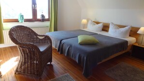 A bedroom with two beds, a wicker chair, a window with curtains, and a nightstand with a lamp.