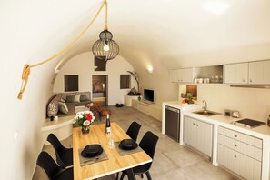 Een moderne keuken met witte kasten, een eethoek met een houten tafel en zwarte stoelen, en een zithoek met een bank en een televisie.