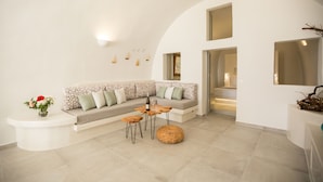 A modern living room with a curved white wall, a sectional sofa with patterned cushions, a wooden coffee table, and a small side table with a vase of flowers. - "Ethereum Luxury Cave" private jacuzzi villa in Imerovigli  (Santorini)