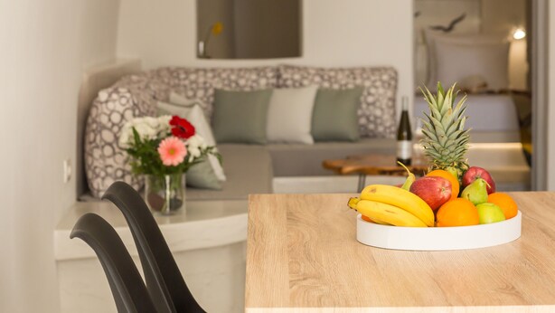 A dining area with a fruit bowl on a wooden table, a sofa with patterned cushions, and a vase with flowers in the background. - "Ethereum Luxury Cave" private jacuzzi villa in Imerovigli  (Santorini)