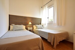 Ein Hotelzimmer mit zwei Einzelbetten, einem Nachttisch mit Lampe, einem Fenster mit Vorhängen und einem querverlaufenden Kopfteil.