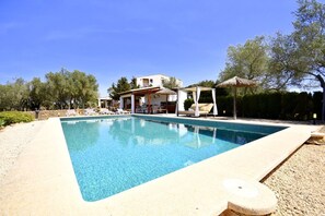 A rectangular swimming pool with clear water, surrounded by a pebble beach, a covered seating area, and a building with a white facade in the background. - 785879 Ses cases - 785879 Ses Cases (Manacor)