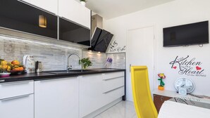 Une cuisine moderne avec des armoires blanches, un comptoir noir et un dosseret carrelé. On y trouve un évier, une machine à café et une plante en pot sur le comptoir. Une chaise jaune est visible, et une horloge murale est accrochée au mur.