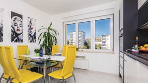 Un coin repas avec des chaises jaunes, une table en verre et une vue sur les bâtiments, visible à travers une grande fenêtre.