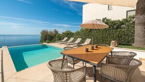 Un espace extérieur avec une piscine, une table en bois et des chaises en osier, offrant une vue sur une mer d’un bleu limpide.