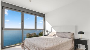 Une chambre dotée d’un grand lit, d’une table de chevet et offrant une vue sur la mer grâce à de grandes fenêtres du sol au plafond.