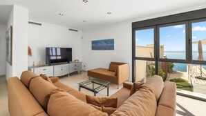 Un salon moderne avec un grand canapé, une table basse, un téléviseur et une vue sur la mer grâce à de grandes fenêtres.