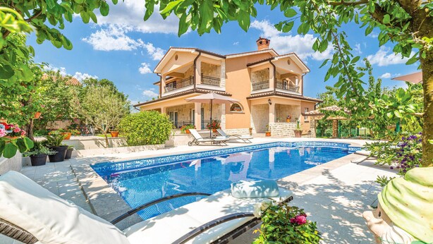 Una casa de dos pisos con piscina, rodeada de plantas en macetas y mobiliario de exterior.