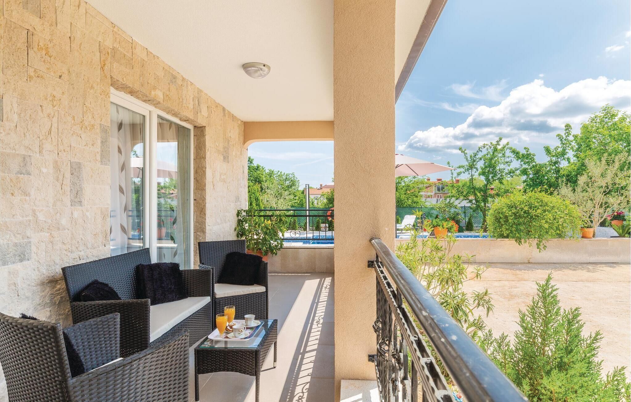 A balcony with wicker furniture, a table with a tray of breakfast items, and a view of a swimming pool and greenery.