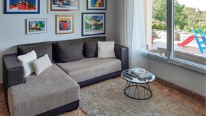 A living room with a sofa, ottoman, and a coffee table. There are framed artworks on the wall and a view of the outdoors through a window.