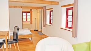 Una habitación con piso de madera, un sofá blanco, un sillón verde, una mesa de comedor con sillas y una ventana con contraventanas rojas.