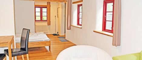 A room with a wooden floor, a white sofa, a green armchair, a dining table with chairs, and a window with red shutters.