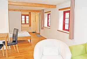 Ein Zimmer mit Holzboden, einem weißen Sofa, einem grünen Sessel, einem Esstisch mit Stühlen und einem Fenster mit roten Lamellen.