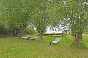 Eine grüne Fläche mit vier Liegestühlen unter Bäumen, einem Picknicktisch und Blick auf ein Gewässer.