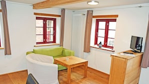 Una habitación con un sofá verde, una mesa de madera, una silla blanca, un escritorio de madera con un televisor y dos ventanas con marcos rojos.