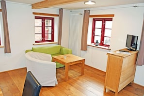 Ein Zimmer mit einem grünen Sofa, einem Holztisch, einem weißen Stuhl, einem Schreibtisch aus Holz mit einem Fernseher und zwei Fenstern mit roten Rahmen.