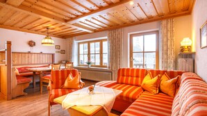 A cozy room with a wooden ceiling, a sofa with orange cushions, a dining table with chairs, and large windows with curtains. - Apartment Rauschberg 67sqm, balcony, kitchenette, 2 bedrooms, 1 living room (Ruhpolding)