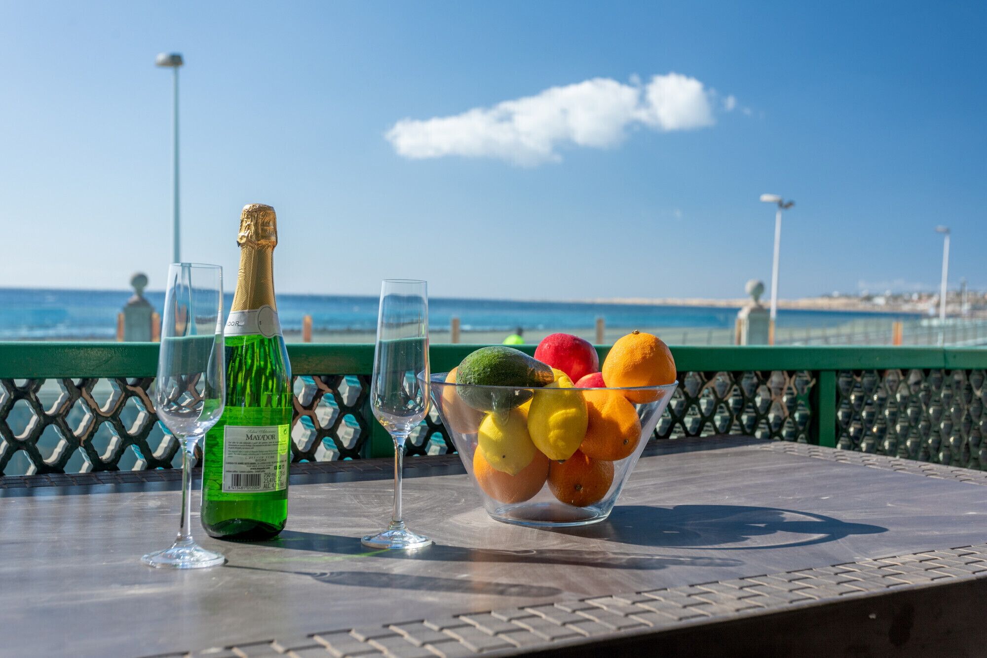 Uma mesa com uma garrafa de champanhe, duas taças e uma cesta de frutas, com vista para o mar.