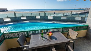 Un área junto a la piscina con una mesa para dos, vista a la playa y una piscina azul cristalina.