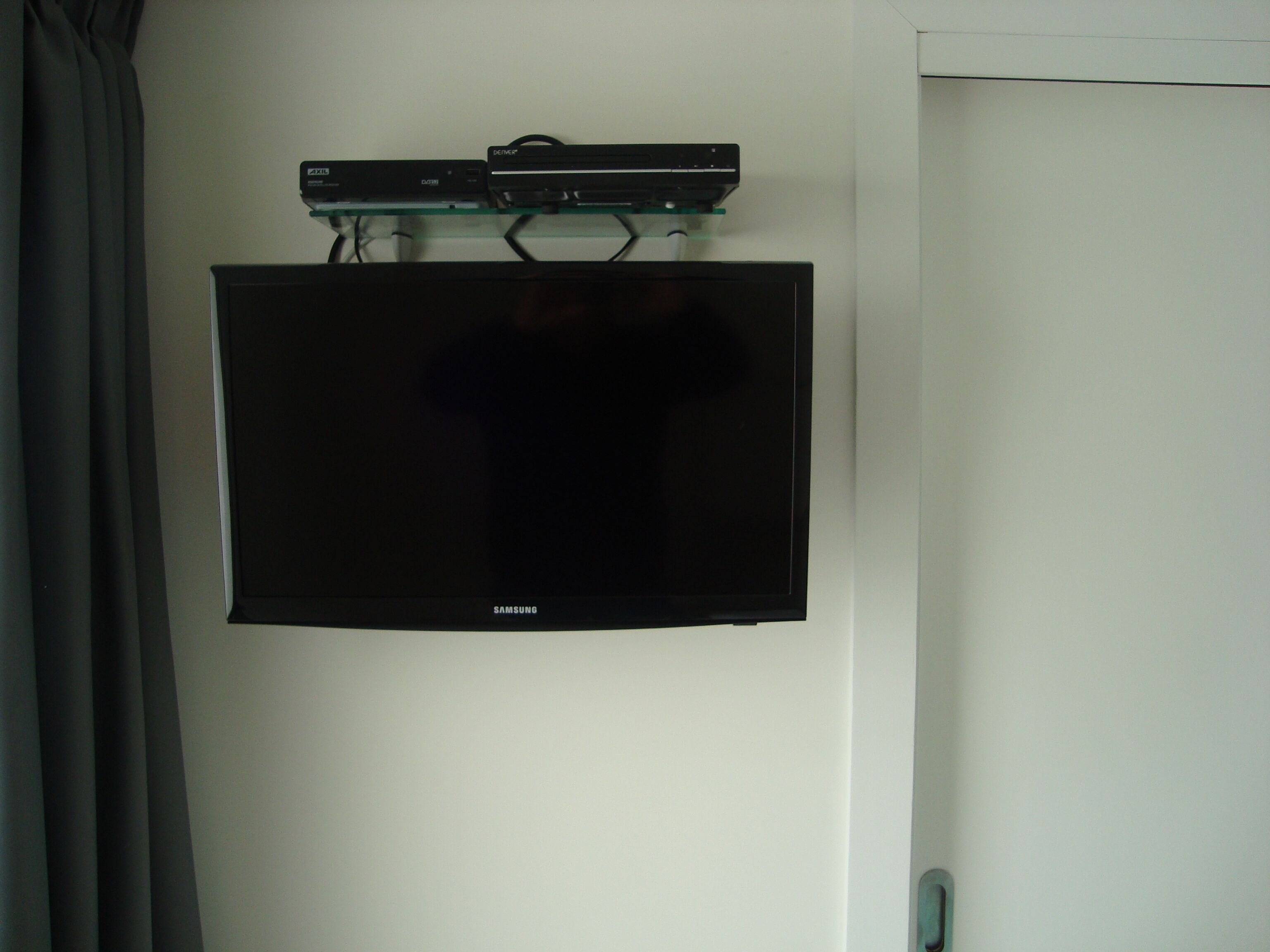 Een Samsung tv bevestigd aan een muur, met een glazen plank erboven waarop een zwart elektronisch apparaat staat.