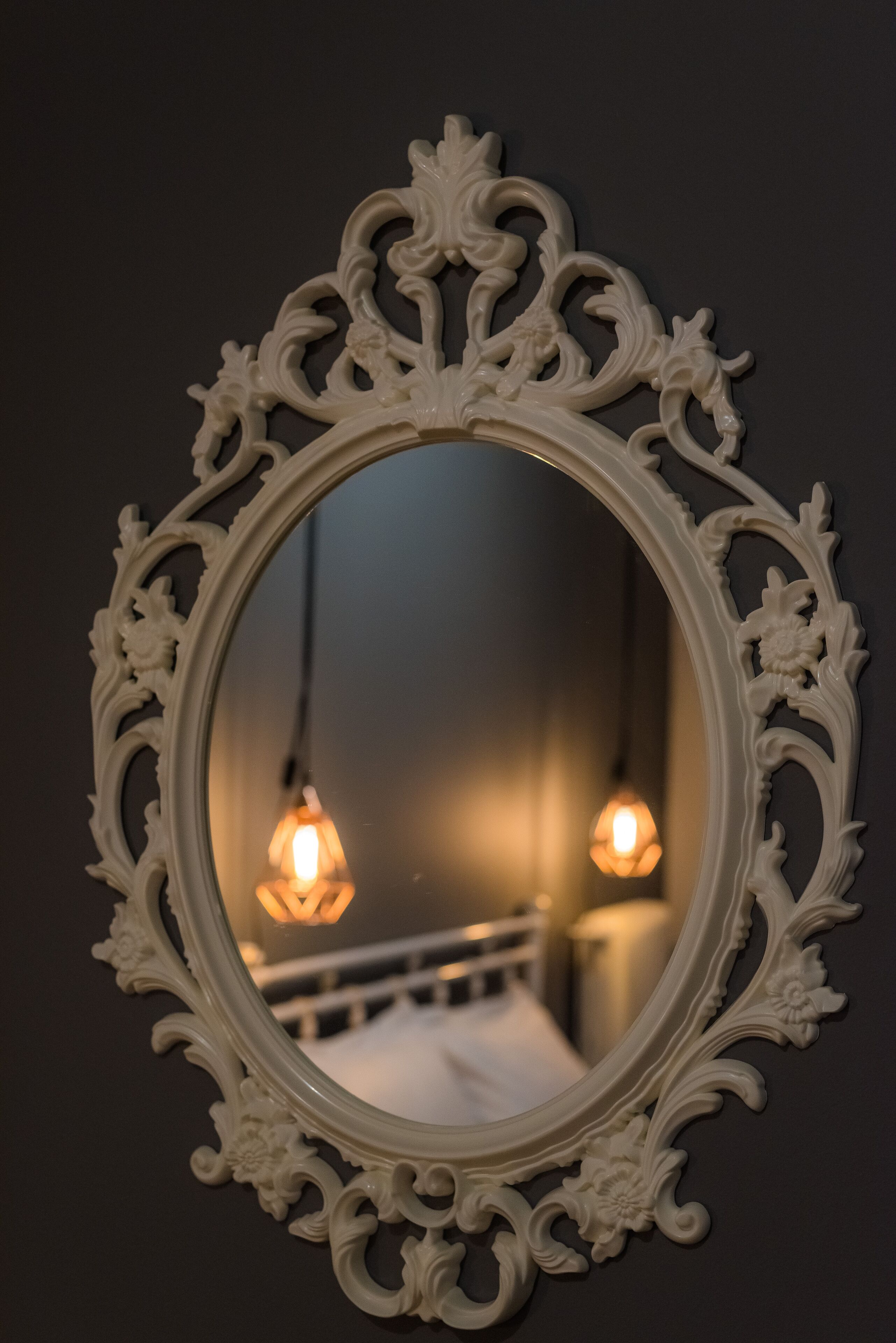 An ornate mirror with a detailed frame reflecting a bed and two hanging light bulbs.
