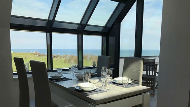 Een eetruimte met een tafel voor vier personen, grote ramen met uitzicht op zee en het landschap, en modern meubilair.