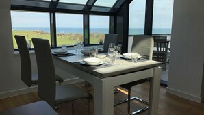 Een eetruimte met een tafel voor vier personen, grote ramen met uitzicht op zee en windturbines, en moderne stoelen.