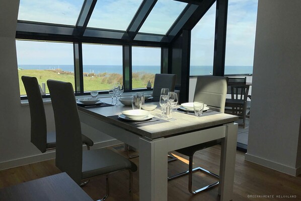 Un coin repas avec une table dressée pour quatre personnes, de grandes fenêtres offrant une vue sur la mer et les éoliennes, et des chaises modernes.